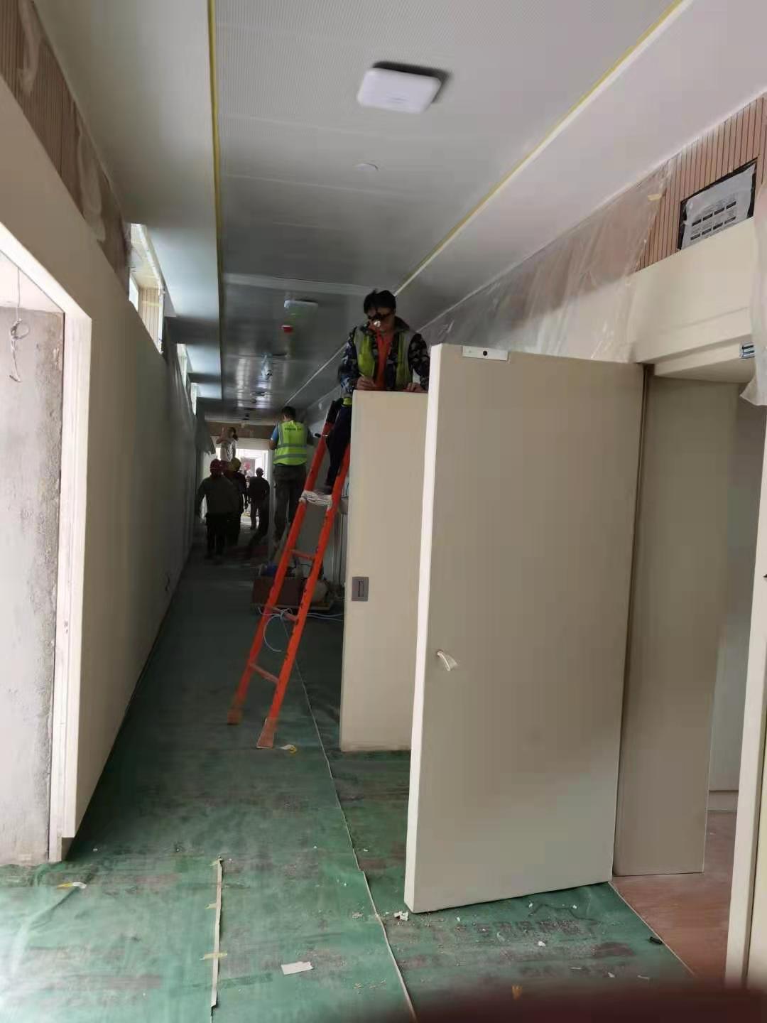 办案区走廊防撞软包墙板安装施工现场，工人攀爬梯子安装吊顶，大面积米色防撞软包板材待安装固定